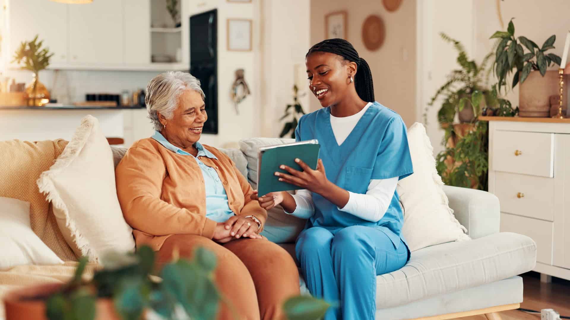 Caregiver supporting aging in place with in-home assistance A senior woman smiling while sitting on a couch with a caregiver in scrubs, reviewing a tablet together in a cozy home setting.